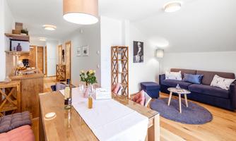 A modern and bright living room with a dining table and a cozy sofa. The decor is stylish and inviting, with decorative elements and wooden shelves.
