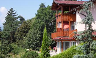 A house with a balcony, surrounded by green trees and bushes. The sky is clear and blue.
