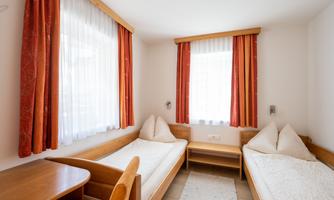 A bright room with two single beds and wooden furniture. Large windows with curtains provide plenty of daylight.