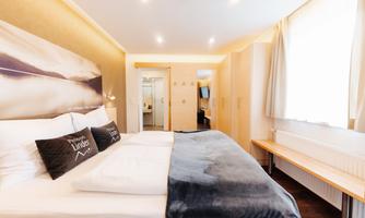 A modern bedroom with a large double bed and bright, inviting decor. The room is well-lit and features a seating area.