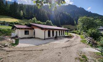 A modern house in a rural setting, surrounded by trees and mountains. The path to the house is paved and the sun is shining.