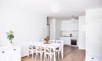 A bright dining room with a white table and chairs. In the background, there is a modern kitchen with white cabinets.