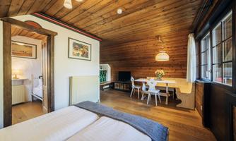 A cozy room with wooden walls and a wooden ceiling. There is a double bed, a table with chairs, and plenty of natural light.