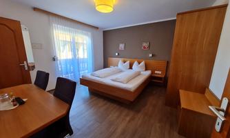 A cozy room with a double bed and wooden furniture. Next to it is a table with chairs and large windows for plenty of light.
