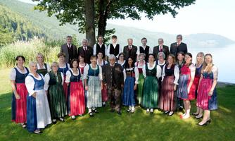 Eine Gruppe von Menschen in traditioneller Tracht steht an einem Seeufer. Im Hintergrund sind Bäume und sanfte Hügel zu sehen.