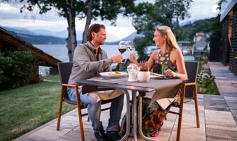 A couple sits smiling at a table outdoors, clinking wine glasses. In the background, there is a beautiful view of a lake and green trees.