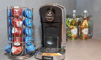 A modern coffee machine stands next to a stand with coffee capsules in various colors. In the background, bottles of syrup are visible.