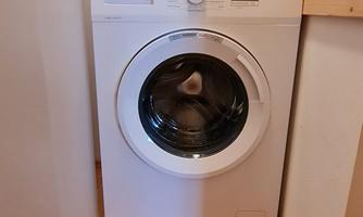 A washing machine is located in a narrow room with a wooden floor. Water connections are visible on the wall.