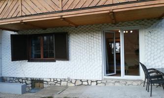 A residential building with a structuring white wall and a wooden veranda. In front of the house are a few chairs and an open window.