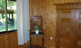 A cozy room with wooden paneling and a large window. There is a bed, a small dresser, and a flower shining in a pot.