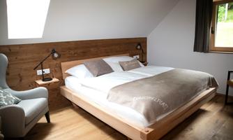 A cozy bedroom with a large bed and wooden decor. Next to it is a comfortable armchair and a window with natural light.