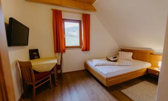 A cozy bedroom with a single bed, a table, and chairs. The room has large windows with orange curtains and a television on the wall.