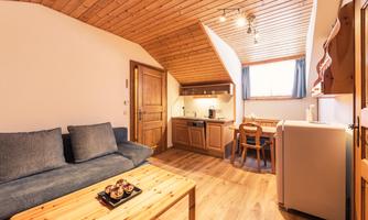 A cozy living space with a sofa corner and a wooden coffee table. In the background, there is a kitchenette and a small dining area.