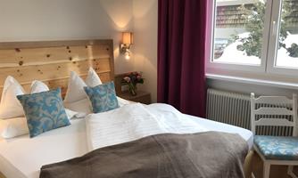 A cozy bedroom with a double bed, bright cushions, and a soft throw. Next to the bed is a chair and a windowsill overlooking a wintery outdoor area.