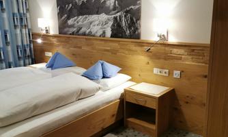 A cozy bedroom with two single beds and blue pillows. A picture of mountains hangs on the wall, and there is a wooden dresser next to the bed.