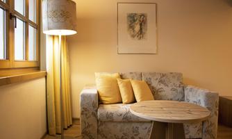 A cozy living room with a sofa and a round wooden table. A lamp light and curtains create a warm atmosphere.
