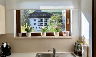 A bright kitchen with a window through which plants can be seen. The view shows a green landscape and a building in the background.