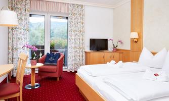 An inviting hotel room with a double bed and a cozy armchair. Large windows offer a view of nature, and the decor is bright and friendly.