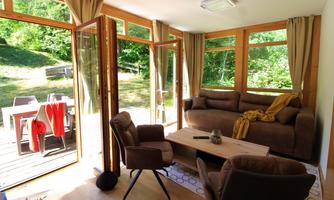 A cozy living area with large windows and plenty of natural light. On the terrace, there are chairs and a table, surrounded by green landscape.
