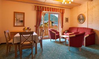 A cozy living room with a red couch and dining table. The room has a balcony with a view of the mountains.