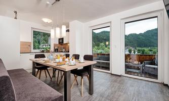 A bright, modern kitchen with a dining area and a table. Large windows offer a view of the mountains and nature outside.