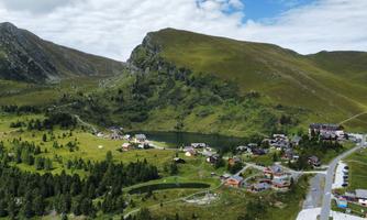 A picturesque mountain village with green meadows and a small lake. In the background, majestic mountains rise under a partly cloudy sky.