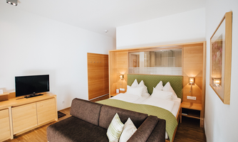 A modern hotel room with a double bed and a cozy seating area. The bright wooden furniture creates an inviting atmosphere.