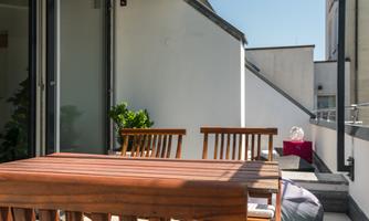 A sunny balcony with a wooden table and chairs. In the background, green plants and a cozy corner can be seen.