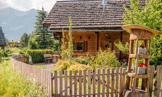 A cozy wooden house with a beautiful garden and a small fence. Surrounded by trees and mountains, the scene exudes a relaxed atmosphere.