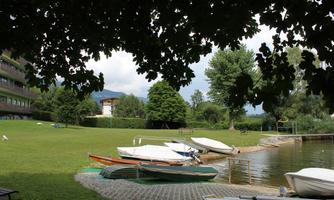 A peaceful lake with boats at the shore, surrounded by green meadow and trees. In the background, some buildings can be seen.