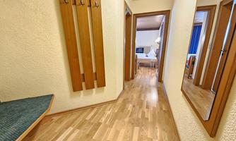 A narrow hallway with a wooden cabinet and a large mirror. The floor is made of wood, and in the background, a bedroom is visible.