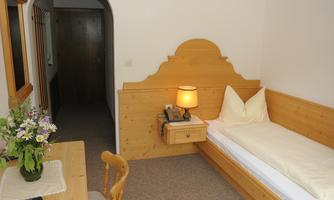 A cozy room with wooden furniture and a single bed.  
On the table, there is a flower vase and a nightstand with a lamp.
