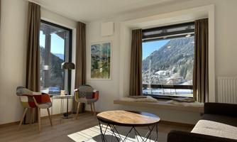 A bright and modern living space with large windows that offer a view of the mountains. There are cozy chairs and a round wooden table.