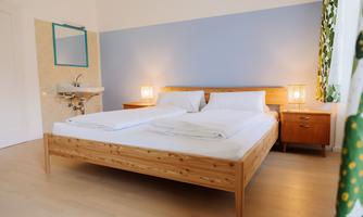 A cozy bedroom with a large wooden bed and bright bedding. The walls are painted in soft blue and earth tones, and there is a small washbasin as well as a window with curtains.