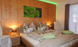 A cozy bedroom with wooden paneling and soft lighting. The double bed is decorated with blooming pillows and a warm blanket.