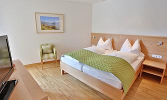 A modern bedroom with a double bed and light wooden furniture. The room is simply and cozily furnished, with a small chair and a television.