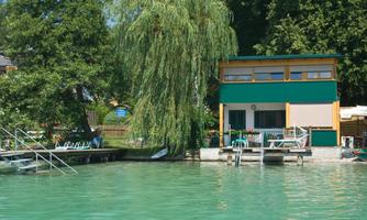 A beautiful house by the water with a green roof and large windows. The surroundings are surrounded by trees, and the water has a clear turquoise color.