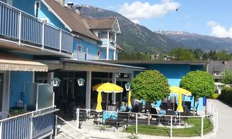 A blue building with a cozy outdoor area. There are sunshades and tables, surrounded by mountains and a clear sky.