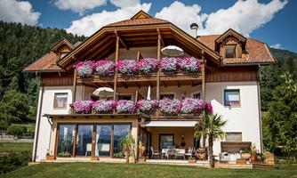 A beautiful house with balconies adorned with colorful flowers. The garden is well-kept and the surroundings are mountainous and green.
