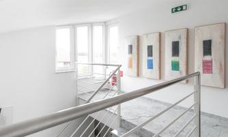 A bright interior with white walls and a modern railing. Several artworks in neutral colors are hanging on the wall.