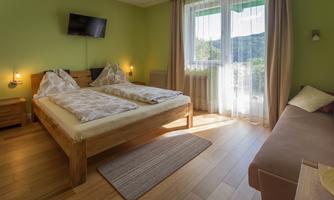 A bright bedroom with two single beds and a large window facing outside. The walls are painted green and there is a small TV on the wall.