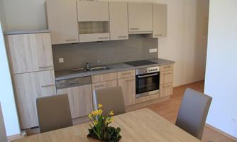 A modern kitchen with light wood surfaces and an elegant dining table. In the background, a dishwasher, a stove, and a sink are visible.