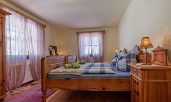 A cozy bedroom with a wooden bed and checkered pillows. The windows are adorned with delicate curtains and there is warm light from a bedside lamp.