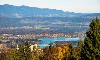 A picturesque landscape with mountains, a clear lake, and colorful trees in autumn. In the foreground, several buildings and forests can be seen.