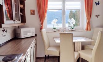 A bright kitchen corner with a small dining table and four chairs. In the background are windows with a view of a landscape and flowers on the windowsill.