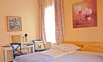 A cozy bedroom with a double bed and soft colors. A painting of a flower hangs on the wall, and a table with a chair is in the room.