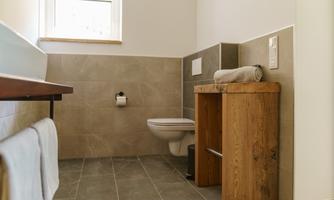 A modern bathroom with a combination of bright and gray tiles. There is a wooden shelf with towels and a toilet.
