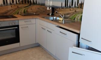 A modern kitchen with white cabinets and a wooden shelf. In the background, a rural landscape with cypress trees is depicted.