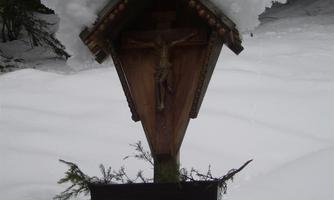 A wooden figure with a crucifix stands against a snowy background. Snow is piled on the roof of the figure.