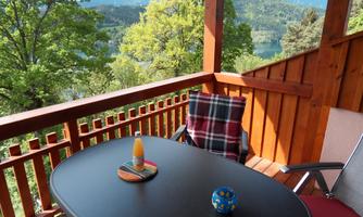 A balcony with a table and chairs, surrounded by trees and overlooking mountains. On the table are decorative items and a colorful drink.